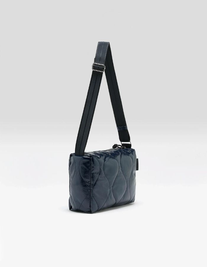 Profil du Sac bandoulière matelassé pour femme en tissu aspect vinyle brillant bleu marine navy avec fermeture zippée et patch en silicone, modèle Hobbes Crossbody Bag de la marque Bellerose.
