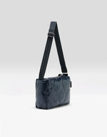 Profil du Sac bandoulière matelassé pour femme en tissu aspect vinyle brillant bleu marine navy avec fermeture zippée et patch en silicone, modèle Hobbes Crossbody Bag de la marque Bellerose.