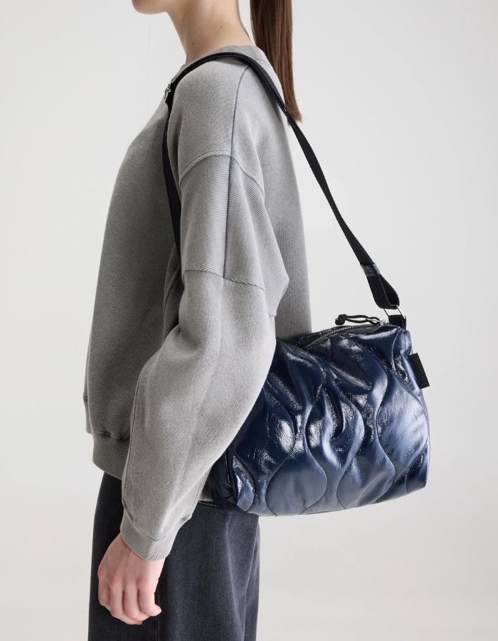 Silhouette avec le Sac bandoulière matelassé pour femme en tissu aspect vinyle brillant bleu marine navy avec fermeture zippée et patch en silicone, modèle Hobbes Crossbody Bag de la marque Bellerose.
