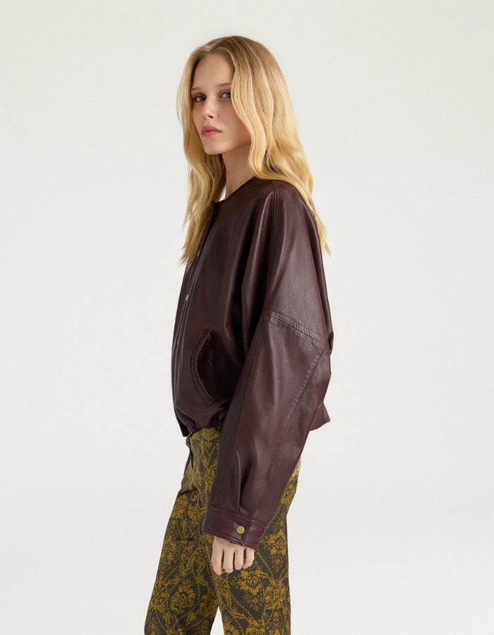 Profil de la Veste bomber courte pour femme en cuir d'agneau marron, modèle Kaldor de la marque MOMONI. Présente une coupe carrée avec cordon de serrage au bas et des boutons-pression en laiton vieilli.
