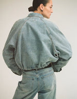 Silhouette de dos avec le Blouson style bomber en jean pour femme de couleur bleu clair délavé Light Blue Denim, modèle Tiana de la marque Rouge Edit. Présente un col chemise évasé, une fermeture à glissière cachée sous des boutons-pression dorés mats Dull Gold et des brides de serrage ajustables.