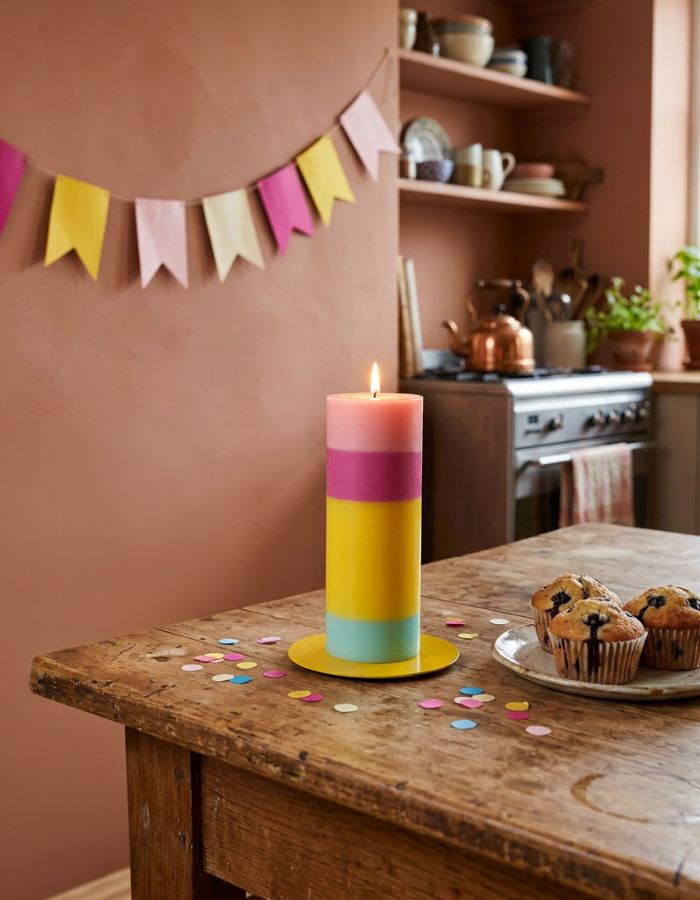 Ambiance déco avec la Grande bougie cylindrique multicolore modèle Hooray Birthday de 20 cm de haut. Confectionnée artisanalement en cire végétale de stéarine biologique avec des couches colorées empilées, présentée à côté de sa boîte en carton naturel.