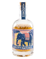 Grande bouteille en verre transparent avec bouchon en liège de la marque Archivist, servant de porte-allumettes. L'étiquette rétro montre un éléphant bleu sur fond de coucher de soleil rose. Le flacon contient de nombreuses allumettes en bois teinté jaune à tête blanche.