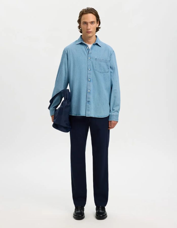 Homme portant une chemise en denim bleu clair Carlo, pantalon marine, et tenant un sac bleu.