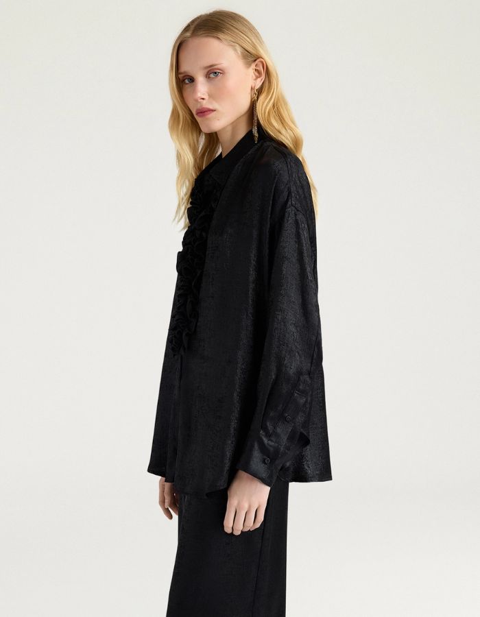Profil du Chemisier long fluide noir pour femme au fini métallisé laminé brillant, modèle Bilancia Bis de la marque Momoni. Présente une coupe oversize avec de grands volants drapés, un col bateau asymétrique tombant sur l'épaule et des manches longues terminées par de hauts poignets à boutons recouverts.