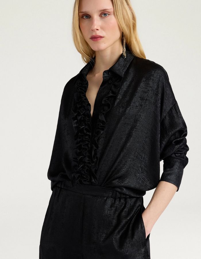 Vue de face du Chemisier long fluide noir pour femme au fini métallisé laminé brillant, modèle Bilancia Bis de la marque Momoni. Présente une coupe oversize avec de grands volants drapés, un col bateau asymétrique tombant sur l'épaule et des manches longues terminées par de hauts poignets à boutons recouverts.