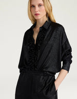 Vue de face du Chemisier long fluide noir pour femme au fini métallisé laminé brillant, modèle Bilancia Bis de la marque Momoni. Présente une coupe oversize avec de grands volants drapés, un col bateau asymétrique tombant sur l'épaule et des manches longues terminées par de hauts poignets à boutons recouverts.
