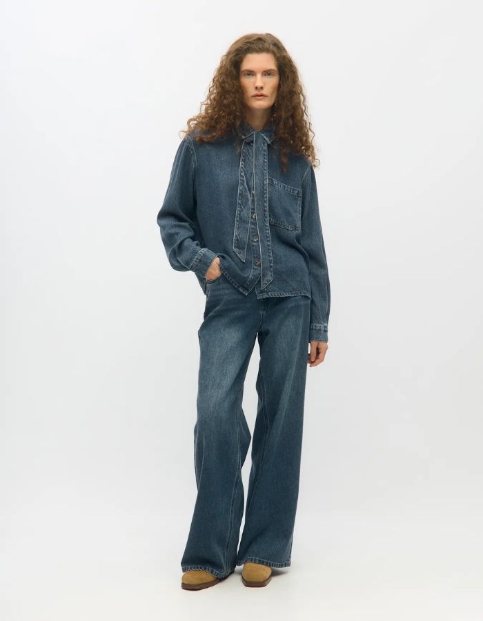 Silhouette avec la Chemise en jean manches longues pour femme de couleur bleu moyen Medium Blue Denim, modèle Rosie de la marque Object. Confectionnée dans une toile de denim souple, elle présente un col chemise, une fermeture boutonnée, une poche plaquée sur la poitrine et un détail de nœud en tissu délicat.
