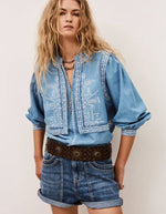 Chemise Laura Ba&sh en denim clair brodé blanc, effet gilet superposé, manches bouffantes, portée avec un short en jean et une ceinture marron.