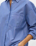 Détail de la Chemise à manches longues pour femme modèle Zila de la marque Object. Tissu tissé fluide à rayures verticales bleu Clématite et blanc, dotée d'un col classique, d'une fermeture boutonnée et d'une poche plaquée sur la poitrine.