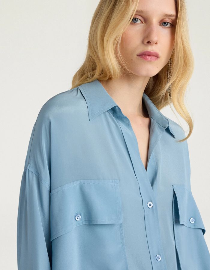Col du Chemisier oversize en pure soie de couleur bleu céruléen uni, modèle Pitanga de la marque Momoni. Présente une coupe ample et fluide avec un col bateau, une fermeture boutonnée originale sur le haut des épaules et des manches longues à poignets à la française.