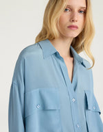 Col du Chemisier oversize en pure soie de couleur bleu céruléen uni, modèle Pitanga de la marque Momoni. Présente une coupe ample et fluide avec un col bateau, une fermeture boutonnée originale sur le haut des épaules et des manches longues à poignets à la française.