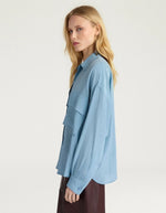 Profil du Chemisier oversize en pure soie de couleur bleu céruléen uni, modèle Pitanga de la marque Momoni. Présente une coupe ample et fluide avec un col bateau, une fermeture boutonnée originale sur le haut des épaules et des manches longues à poignets à la française.