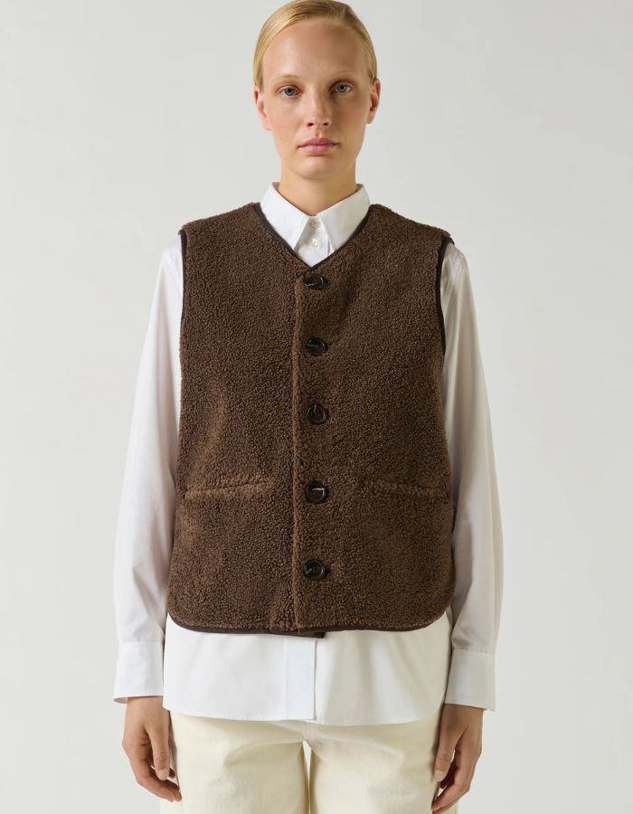 Vue de face du Gilet sans manches pour femme en fausse fourrure de couleur marron foncé, modèle Fusil de la marque Soeur. Présente une coupe droite dotée d'une encolure en V, d'une fermeture boutonnée à l'avant, de deux poches passepoilées et de bordures proprement gansées.
