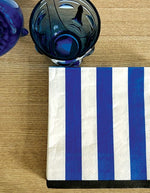 Vue à plat des serviettes de table carrées en papier recyclé de 33 cm de côté, décorées d'un motif graphique imprimé de rayures bleues et blanches alternées.