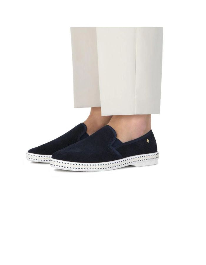 Allure des Mocassins Rivieras Classic en daim bleu marine perforé. Semelle blanche contrastée avec couture selle en coton, élastiques de confort et rivet métallique logoté sur le côté. Design intemporel fabriqué en Espagne.