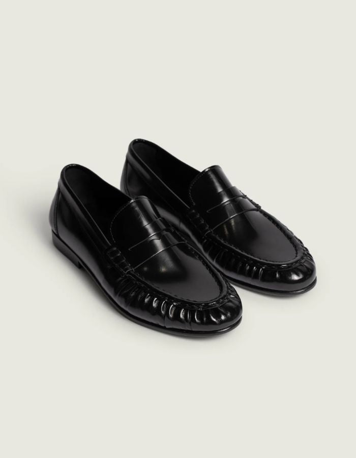 Profil des Mocassins en cuir noir brillant pour homme et femme de type Penny Loafers, modèle Wrinkle de la marque POSA. Chaussures basses en cuir italien box poli avec une bride classique sur le coup de pied, un bout arrondi à empiècement plissé texturé et une semelle noire fine.
