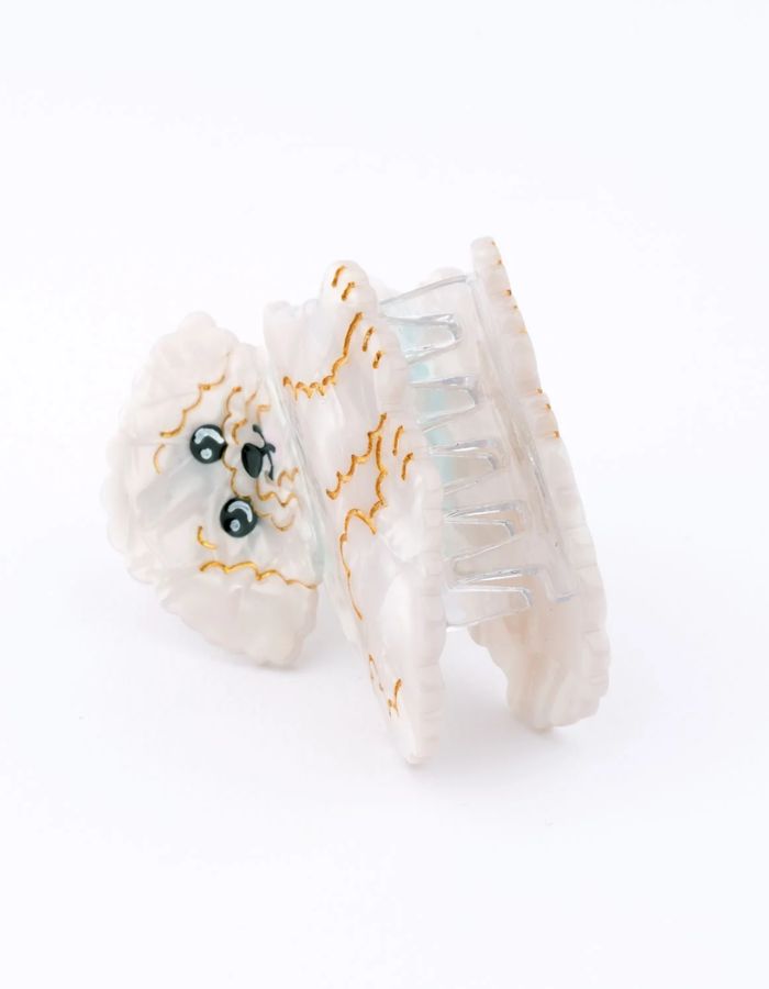 Gros plan sur la Petite pince à cheveux crabe de la marque Coucou Suzette en acétate de cellulose blanc nacré brillant. L'accessoire prend la forme d'un chien bichon frisé dodu portant un collier bleu turquoise avec des détails dorés.