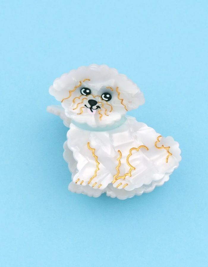 Petite pince à cheveux crabe de la marque Coucou Suzette en acétate de cellulose blanc nacré brillant. L'accessoire prend la forme d'un chien bichon frisé dodu portant un collier bleu turquoise avec des détails dorés.