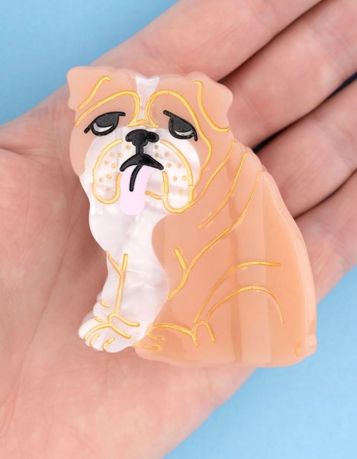 Gros plan sur la Petite pince à cheveux crabe de la marque Coucou Suzette en acétate de cellulose brillant. L'accessoire représente un chien bouledogue anglais marron et blanc de profil avec de jolis bourrelets, des petits yeux noirs et une petite langue rose pendante.