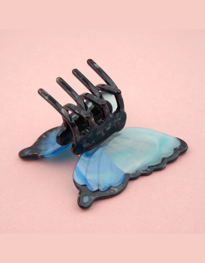 Gros plan sur la Pince à cheveux crabe de taille moyenne de la marque Coucou Suzette en acétate de cellulose brillant. L'accessoire prend la forme d'un papillon bleu azur et blanc nacré aux ailes déployées avec des contours noirs.