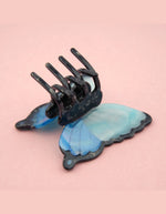 Gros plan sur la Pince à cheveux crabe de taille moyenne de la marque Coucou Suzette en acétate de cellulose brillant. L'accessoire prend la forme d'un papillon bleu azur et blanc nacré aux ailes déployées avec des contours noirs.