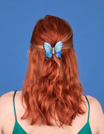 Mise en situation de la Pince à cheveux crabe de taille moyenne de la marque Coucou Suzette en acétate de cellulose brillant. L'accessoire prend la forme d'un papillon bleu azur et blanc nacré aux ailes déployées avec des contours noirs.