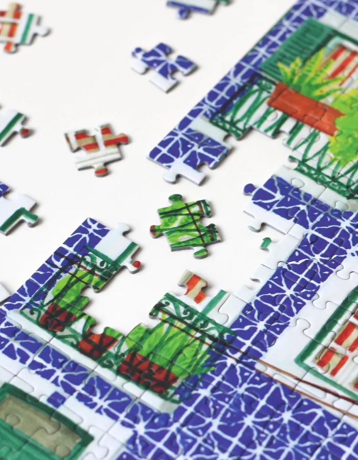 Détail du Puzzle de 1000 pièces pour adulte illustré par Mélanie Voituriez pour Piecely, représentant une rue colorée de Lisbonne avec le tramway jaune traditionnel et des façades en azulejos. Présenté aux côtés de sa boîte de rangement écoresponsable.