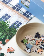 Détail de la Boîte de puzzle 1000 pièces Paris de la marque Piecely. L'illustration représente une table de petit-déjeuner sur un balcon avec croissants et café, offrant une vue romantique sur les toits de Paris et la Tour Eiffel au lever du soleil.