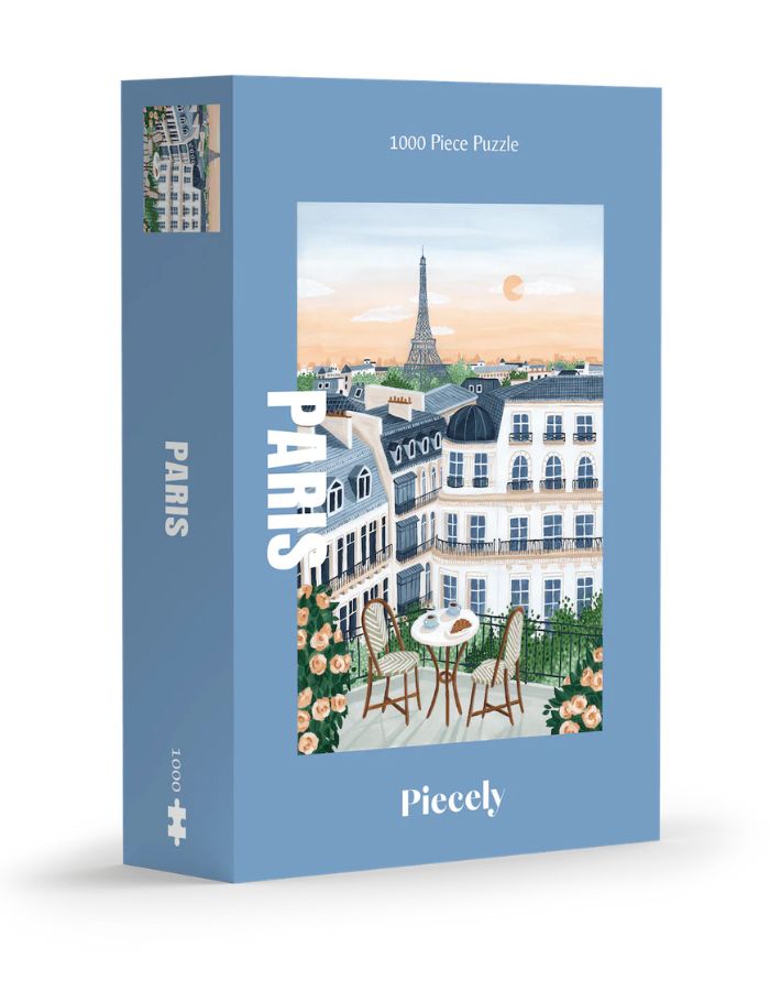 Boîte de puzzle 1000 pièces Paris de la marque Piecely. L'illustration représente une table de petit-déjeuner sur un balcon avec croissants et café, offrant une vue romantique sur les toits de Paris et la Tour Eiffel au lever du soleil.