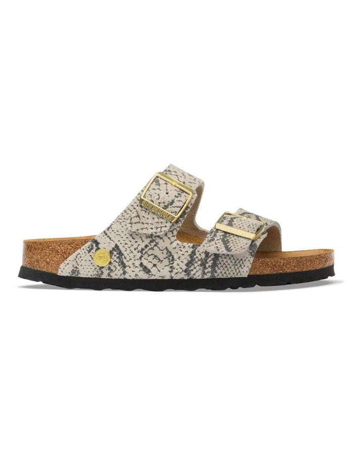 Profil des Sandales pour pieds fins en largeur Narrow, modèle emblématique Arizona Rivet Logo de la marque Birkenstock. Confectionnées en cuir velours souple de couleur claire beige huître Snake Oyster, elles présentent deux brides horizontales ajustables avec boucles en métal, un rivet logo et la célèbre semelle anatomique marron en liège naturel
