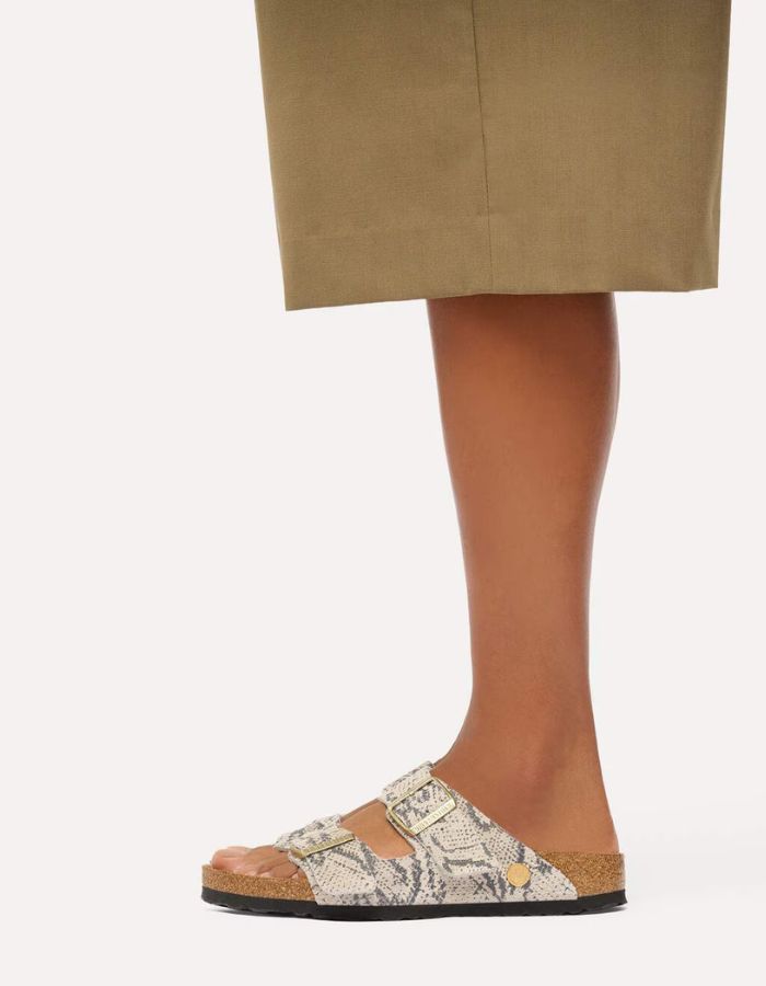 Silhouette avec les Sandales pour pieds fins en largeur Narrow, modèle emblématique Arizona Rivet Logo de la marque Birkenstock. Confectionnées en cuir velours souple de couleur claire beige huître Snake Oyster, elles présentent deux brides horizontales ajustables avec boucles en métal, un rivet logo et la célèbre semelle anatomique marron en liège naturel