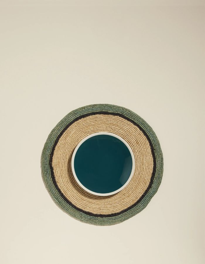 Set de table rond en raphia tressé Naturel et Céladon avec bordure foncée, sous une assiette bleue.