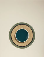 Set de table rond en raphia tressé Naturel et Céladon avec bordure foncée, sous une assiette bleue.
