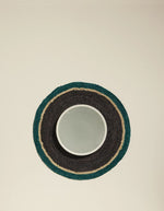 Set de table rond en raphia tressé main, noir, bleu Sarah et beige, avec un bol blanc.