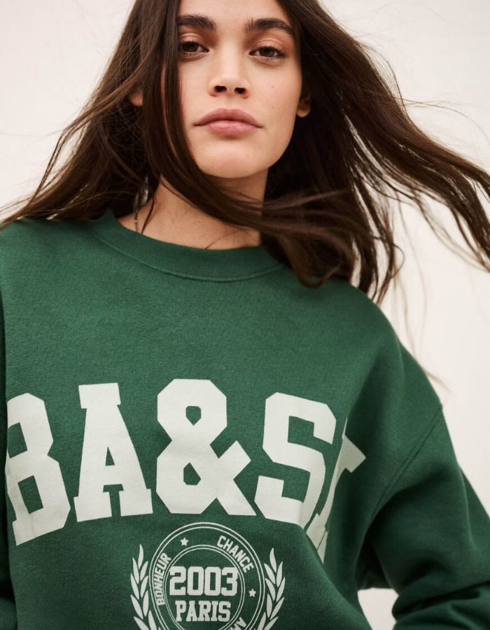 Femme portant un sweatshirt Fenja Emeraude Ba&sh vert foncé avec logo "BA&SH" blanc et motif campus.