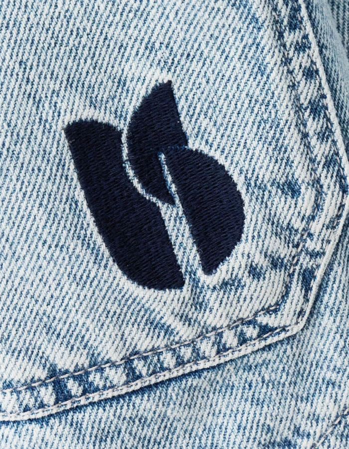 Logo brodé bleu marine sur la poche poitrine d'une veste en jean bleu clair délavé.