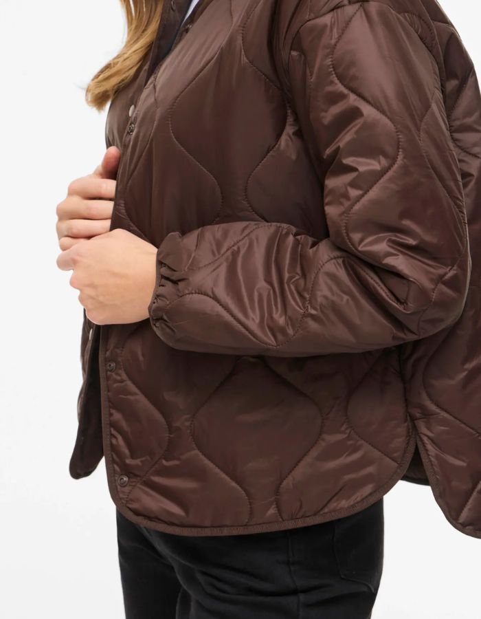 Manche de la Veste matelassée légère et courte pour femme de couleur marron chocolat, modèle Dune de la marque Vila. Présente un col en V, des manches longues, des poches plaquées à l'avant et une fermeture par boutons-pression.
