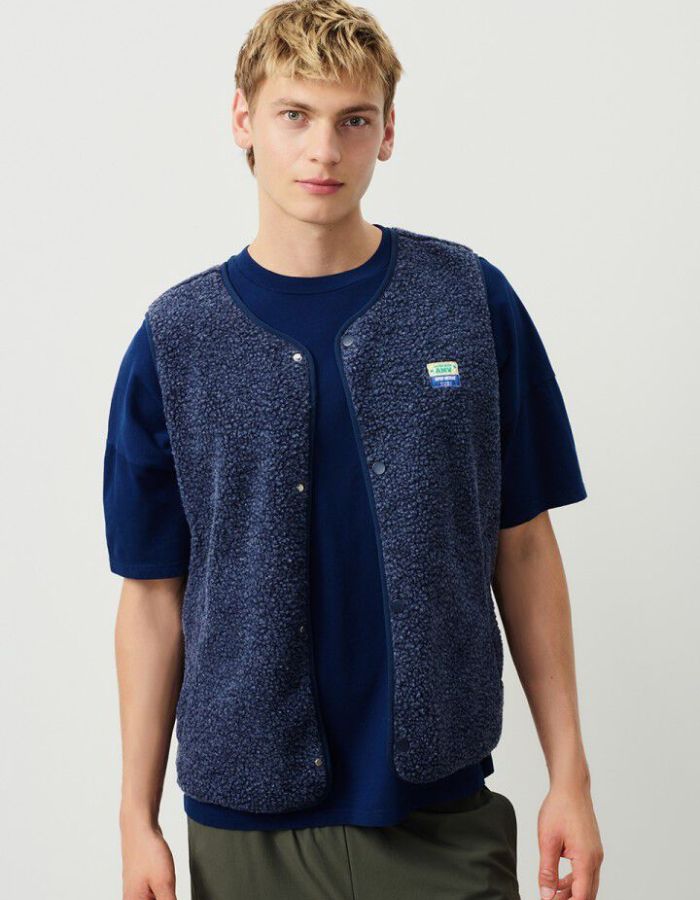 Vue de face de la Veste sans manches pour homme de couleur bleu nuit foncé Astre Chiné, modèle Hoktown de la marque American Vintage. Confectionnée dans une matière épaisse effet mouton retourné shearling, elle présente un col montant bas, une fermeture à boutons-pression, deux poches zippées et un patch logo sur la poitrine.