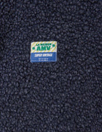 Logo de la Veste sans manches pour homme de couleur bleu nuit foncé Astre Chiné, modèle Hoktown de la marque American Vintage. Confectionnée dans une matière épaisse effet mouton retourné shearling, elle présente un col montant bas, une fermeture à boutons-pression, deux poches zippées et un patch logo sur la poitrine.