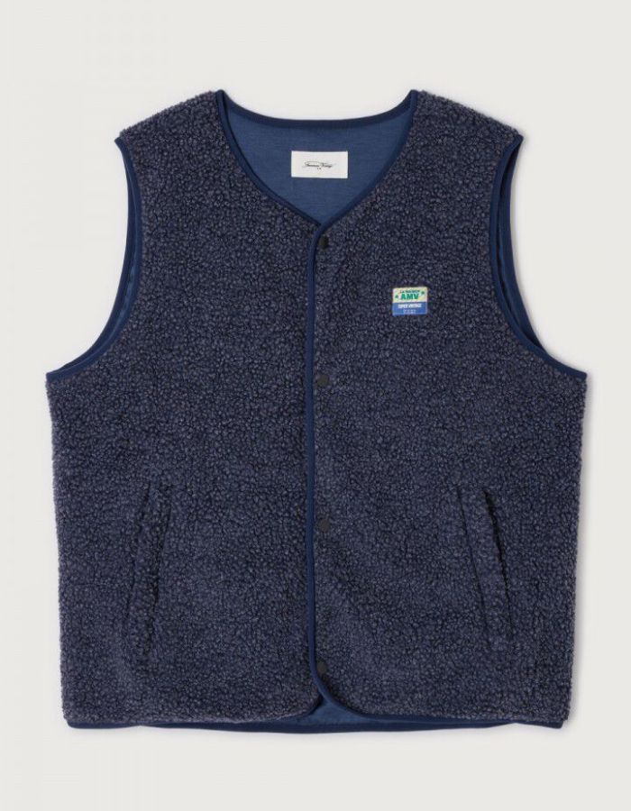 Veste sans manches pour homme de couleur bleu nuit foncé Astre Chiné, modèle Hoktown de la marque American Vintage. Confectionnée dans une matière épaisse effet mouton retourné shearling, elle présente un col montant bas, une fermeture à boutons-pression, deux poches zippées et un patch logo sur la poitrine.