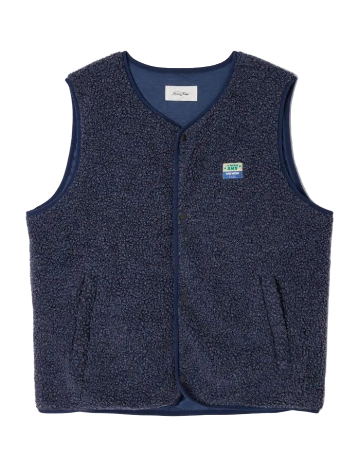 Veste sans manches pour homme de couleur bleu nuit foncé Astre Chiné, modèle Hoktown de la marque American Vintage. Confectionnée dans une matière épaisse effet mouton retourné shearling, elle présente un col montant bas, une fermeture à boutons-pression, deux poches zippées et un patch logo sur la poitrine.