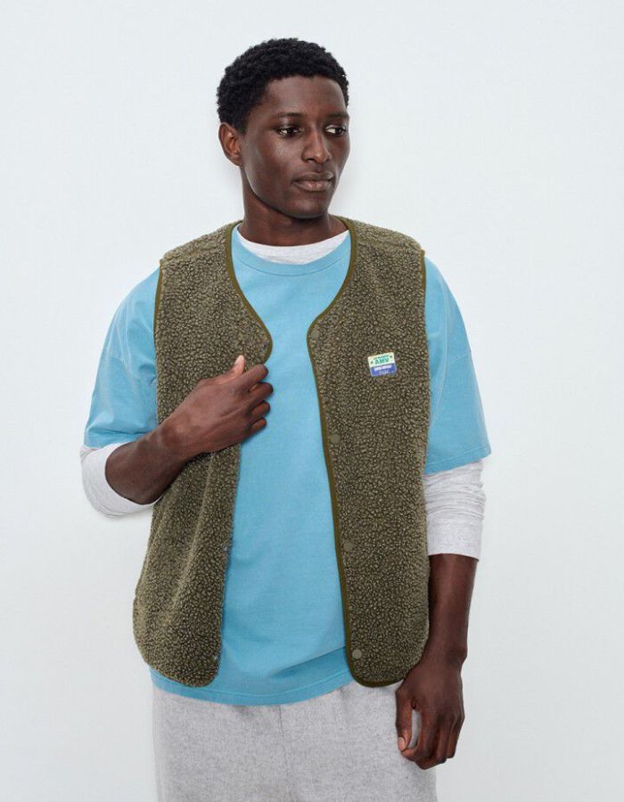 Vue de face de la Veste sans manches pour homme de couleur vert kaki Varan Chiné, modèle Hoktown de la marque American Vintage. Confectionnée dans une matière épaisse effet mouton retourné shearling, elle présente un col montant bas, une fermeture à boutons-pression, deux poches zippées et un patch logo sur la poitrine.
