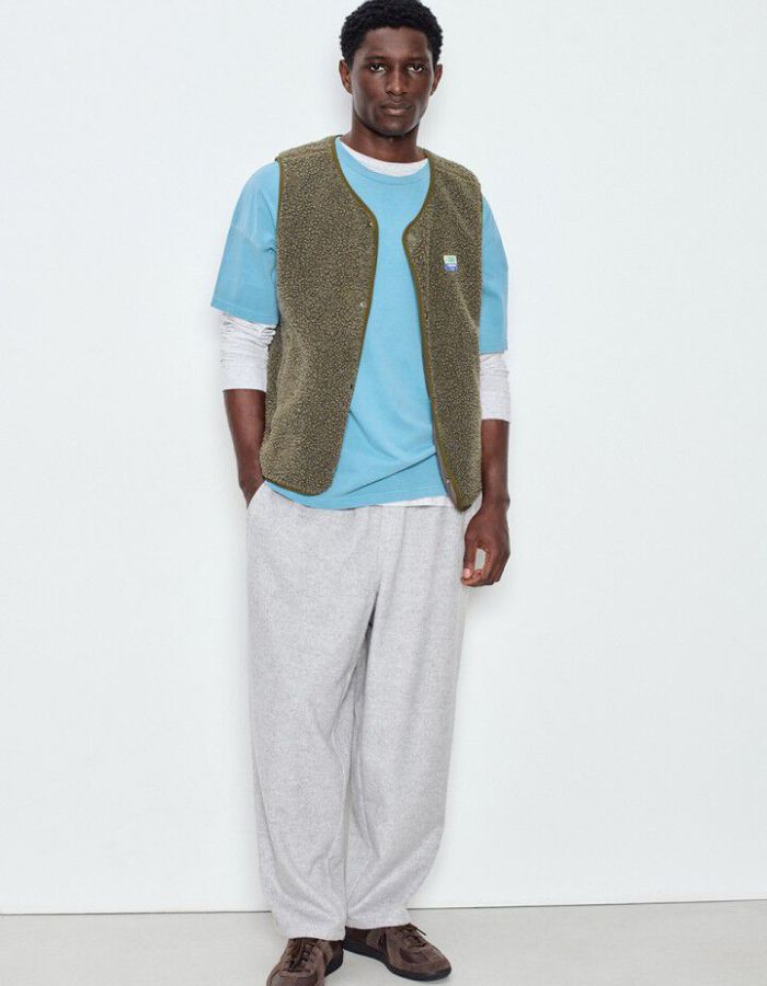 Silhouette avec la Veste sans manches pour homme de couleur vert kaki Varan Chiné, modèle Hoktown de la marque American Vintage. Confectionnée dans une matière épaisse effet mouton retourné shearling, elle présente un col montant bas, une fermeture à boutons-pression, deux poches zippées et un patch logo sur la poitrine.
