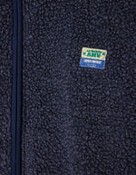 Logo de la Veste zippée à manches longues pour homme de couleur bleu nuit foncé Astre Chiné, modèle Hoktown de la marque American Vintage. Confectionnée dans une matière épaisse effet mouton retourné shearling, elle présente un col montant bas, une fermeture éclair, deux poches zippées et un écusson logo AMV sur la poitrine.