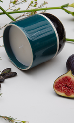Bougie Passy Figue Maison Sarah Lavoine en céramique bicolore bleu canard et noir, avec une mèche blanche, entourée de feuilles et de figues fraîches.