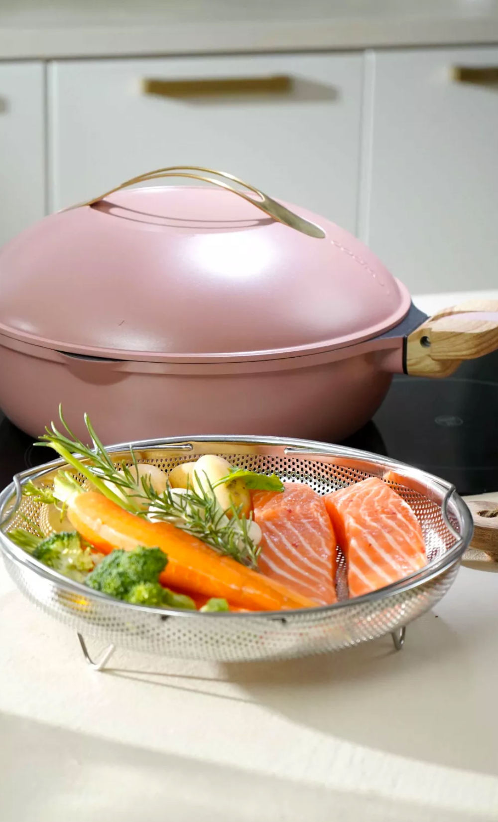 La Fabuleuse Poêle Cookut rose 28cm avec couvercle doré, et un panier vapeur en métal rempli de saumon et légumes.