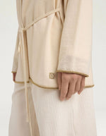Manche du Cardigan à col rond pour femme en pure laine crème, modèle Klak Bis de la marque MOMONI. Présente des manches raglan amples, une fermeture à ruban de laine à nouer et des liserés en lurex doré.