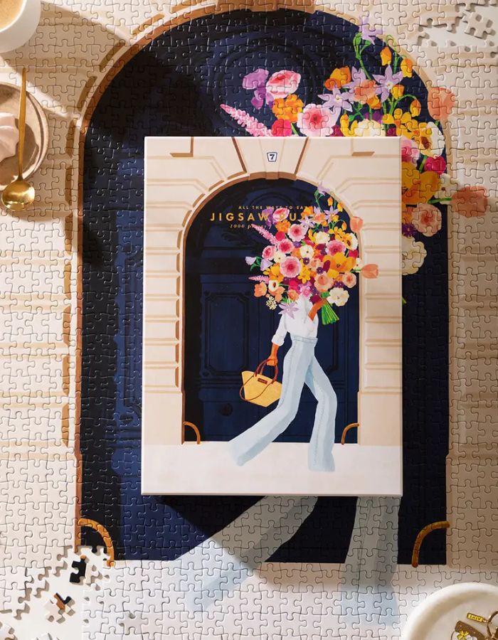 Ambiance avec le puzzle assemblé de 1000 pièces Parisian Door par All The Ways To Say montrant une femme avec un bouquet de fleurs devant une porte cochère bleue à Paris