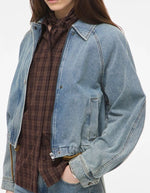 Devant du Blouson style bomber en jean pour femme de couleur bleu clair délavé Light Blue Denim, modèle Tiana de la marque Rouge Edit. Présente un col chemise évasé, une fermeture à glissière cachée sous des boutons-pression dorés mats Dull Gold et des brides de serrage ajustables.